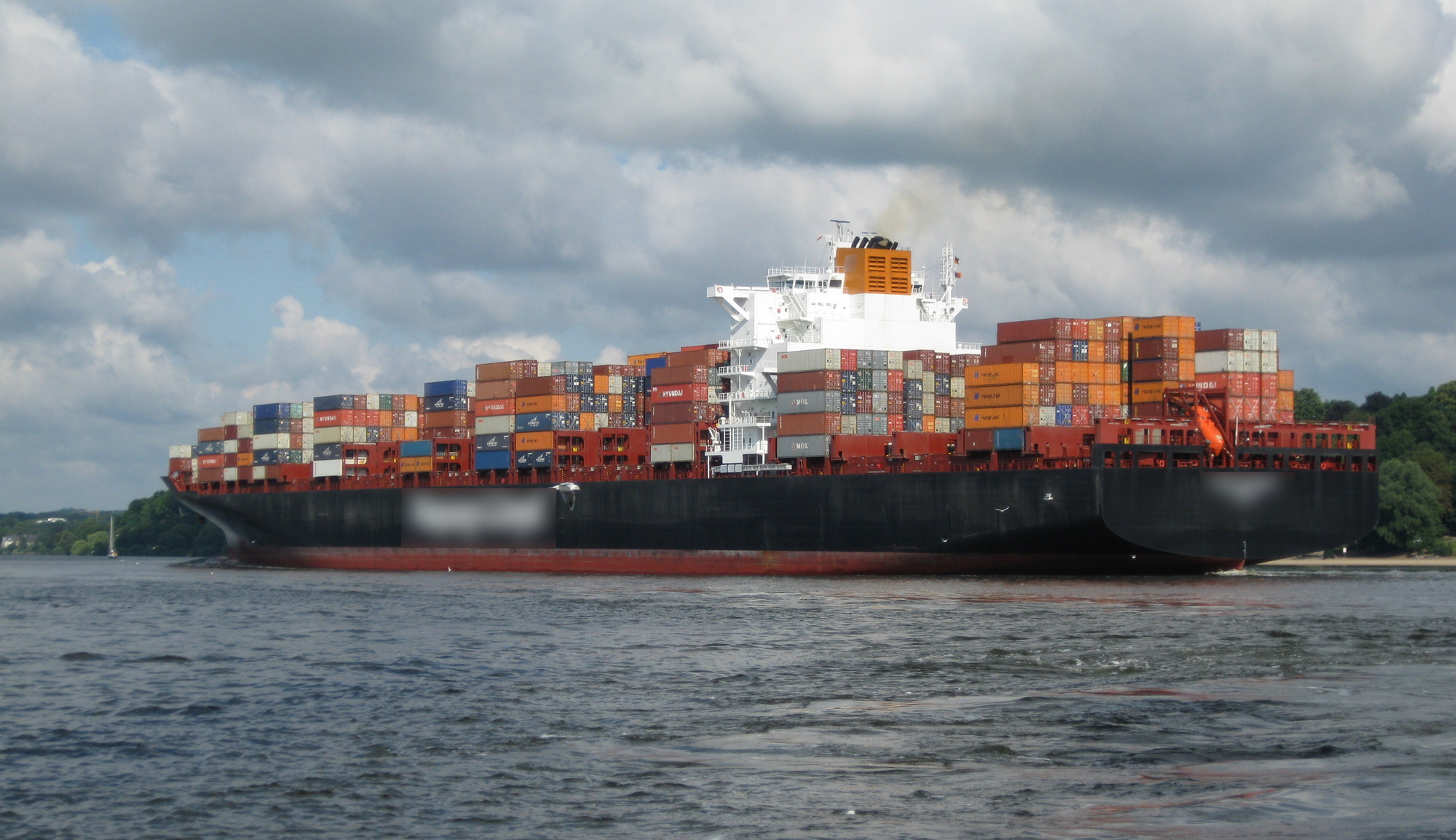 Containerfrachter auf der Elbe Ein großes Frachtschiff, beladen mit zahlreichen gestapelten Frachtcontainern, fährt unter einem bewölkten Himmel auf dem Wasser. Das Schiff ist schwarz mit einem weißen Überbau und zeigt bunte Container in verschiedenen Blau-, Rot-, Orange- und Gelbtönen. Im Hintergrund sind Bäume zu sehen.