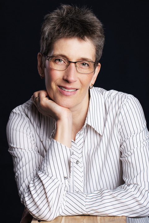 Eine Person mit kurzen grauen Haaren und Brille lächelt und stützt den Kopf auf die Hand. Sie trägt ein weiß gestreiftes Hemd und sitzt vor einem dunklen Hintergrund. Sie macht vielleicht eine Pause von ihrer Meisterarbeit-Korrektur. Portraitfoto von Sabine